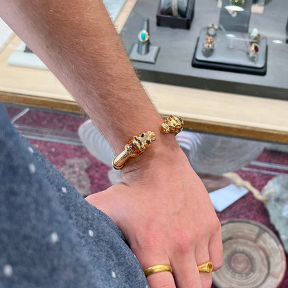 A 14 karat yellow gold hinged bangle bracelet featuring the motif of two panther heads.  There are yellow and red accent stones.