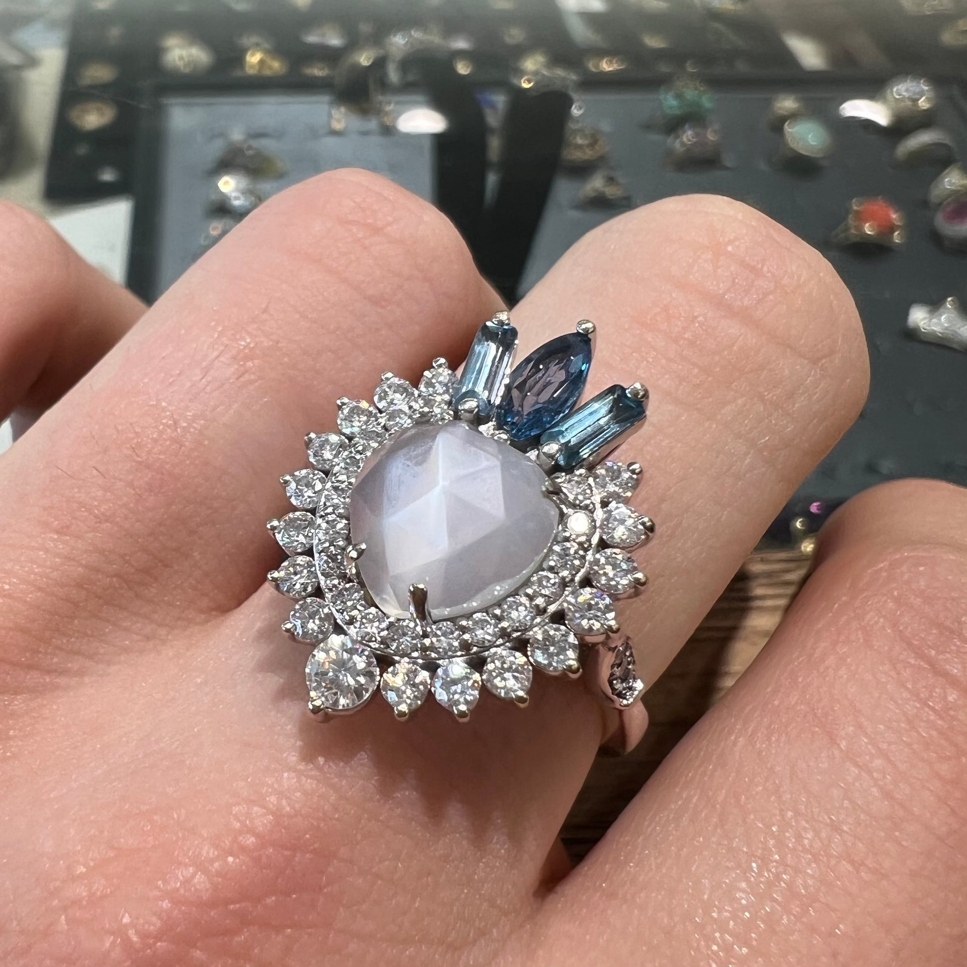 A ladies' white gold ring set with a faceted, heart shaped white moonstone, round moissanites, two aquamarine baguettes, and a marquise cut blue sapphire.