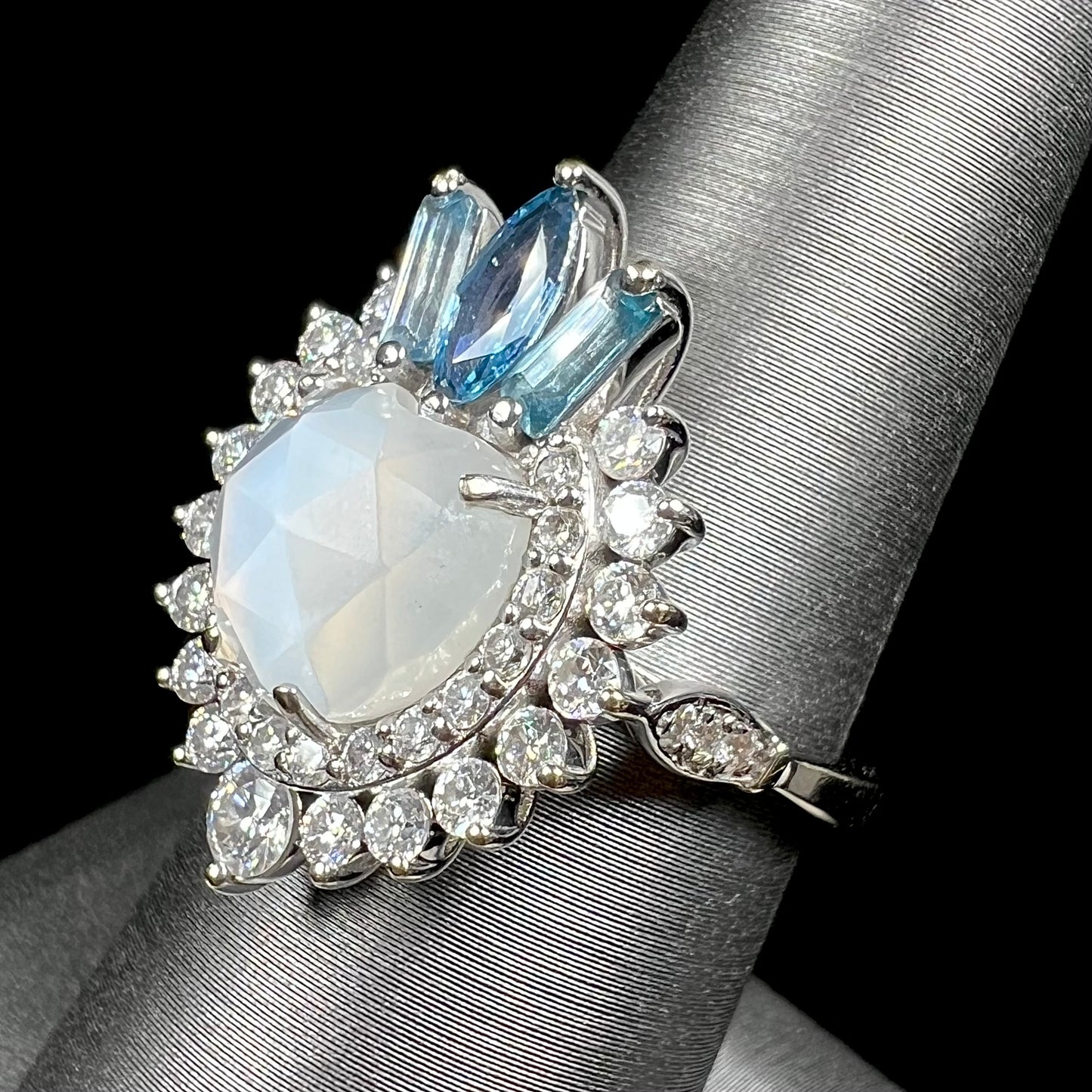 A ladies' white gold ring set with a faceted, heart shaped white moonstone, round moissanites, two aquamarine baguettes, and a marquise cut blue sapphire.