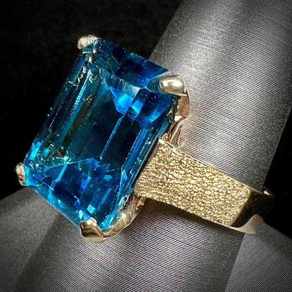 A ladies' yellow gold London blue topaz solitaire ring.  The ring is textured, and the topaz is emerald cut.