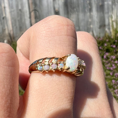 A yellow gold ring featuring two crossing, alternating lines of round cut synthetic opals.  In the middle is a larger oval cut synthetic opal.