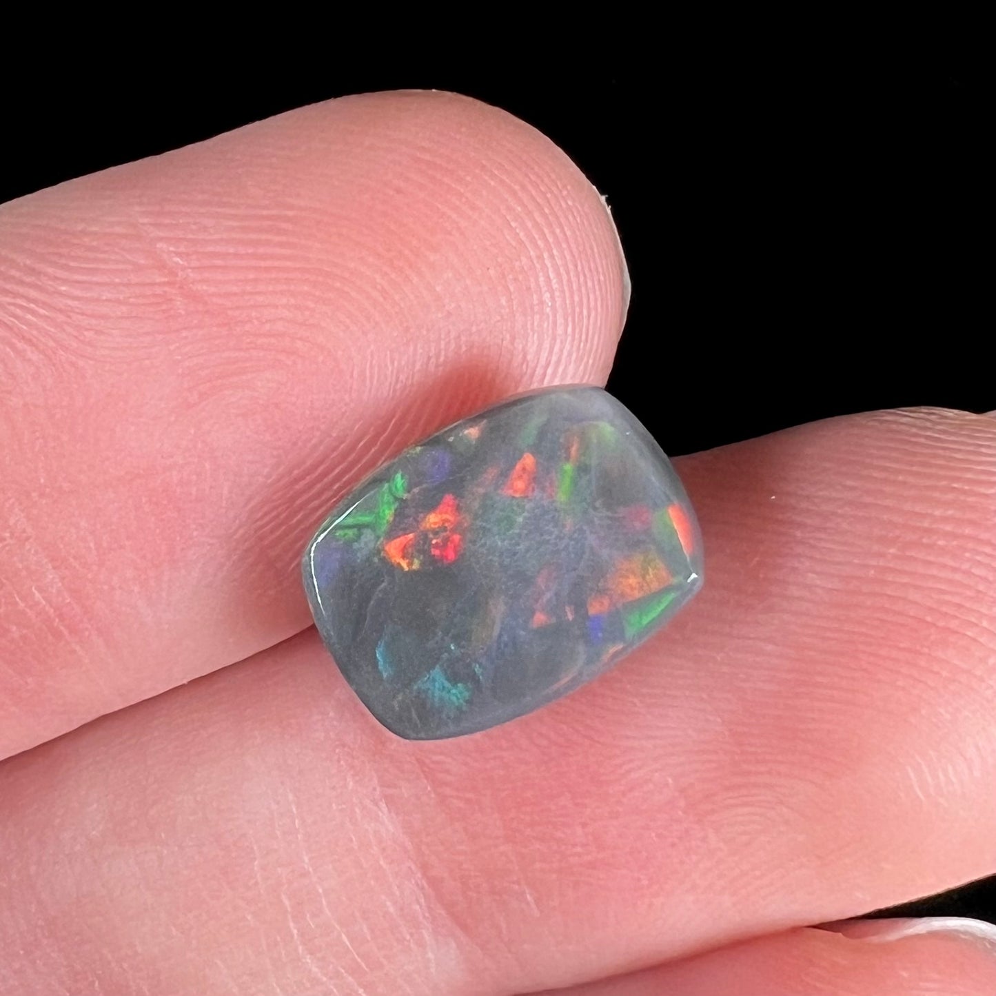 A loose, cushion cut black opal stone.  The stone plays colors of red, green, blue, and purple.