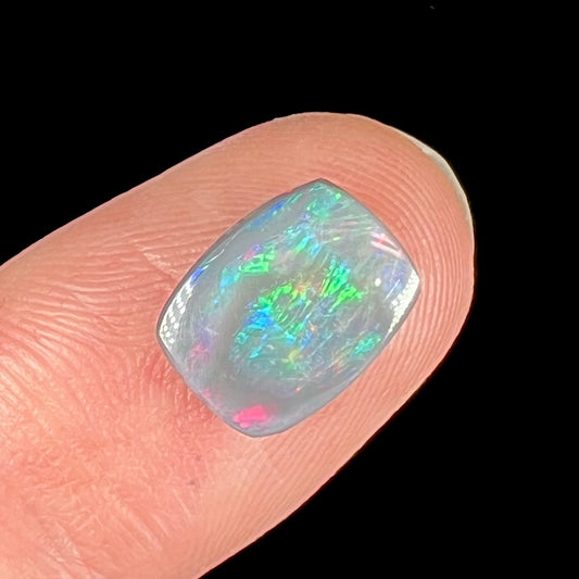 A loose, cushion cabochon cut semi-black opal stone.  The stone plays multiple colors.