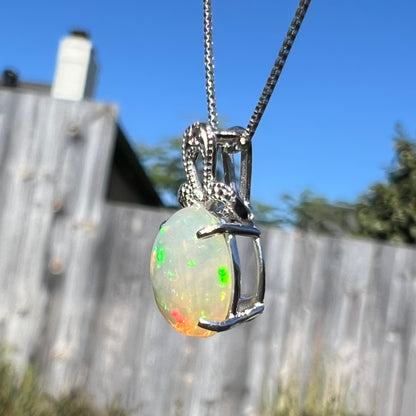 An oval cut Ethiopian opal mounted in a diamond-accented, 18 karat white gold pendant on a box chain.