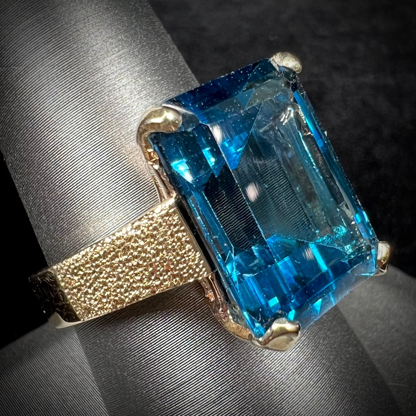 A ladies' yellow gold London blue topaz solitaire ring.  The ring is textured, and the topaz is emerald cut.