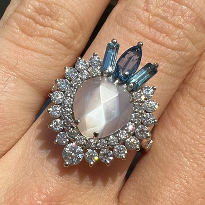 A ladies' white gold ring set with a faceted, heart shaped white moonstone, round moissanites, two aquamarine baguettes, and a marquise cut blue sapphire.