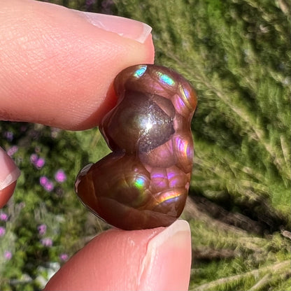 A polished, freeform shaped fire agate stone.  The stone is purple, green, and blue with red and yellow banding.