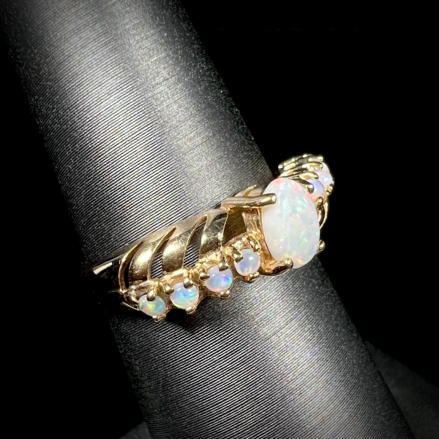 A yellow gold ring featuring two crossing, alternating lines of round cut synthetic opals.  In the middle is a larger oval cut synthetic opal.