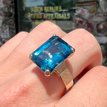 A ladies' yellow gold London blue topaz solitaire ring.  The ring is textured, and the topaz is emerald cut.