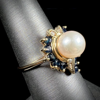 A yellow gold, heart shaped ring set with a freshwater pearl surrounded by blue sapphire and diamond accent stones.