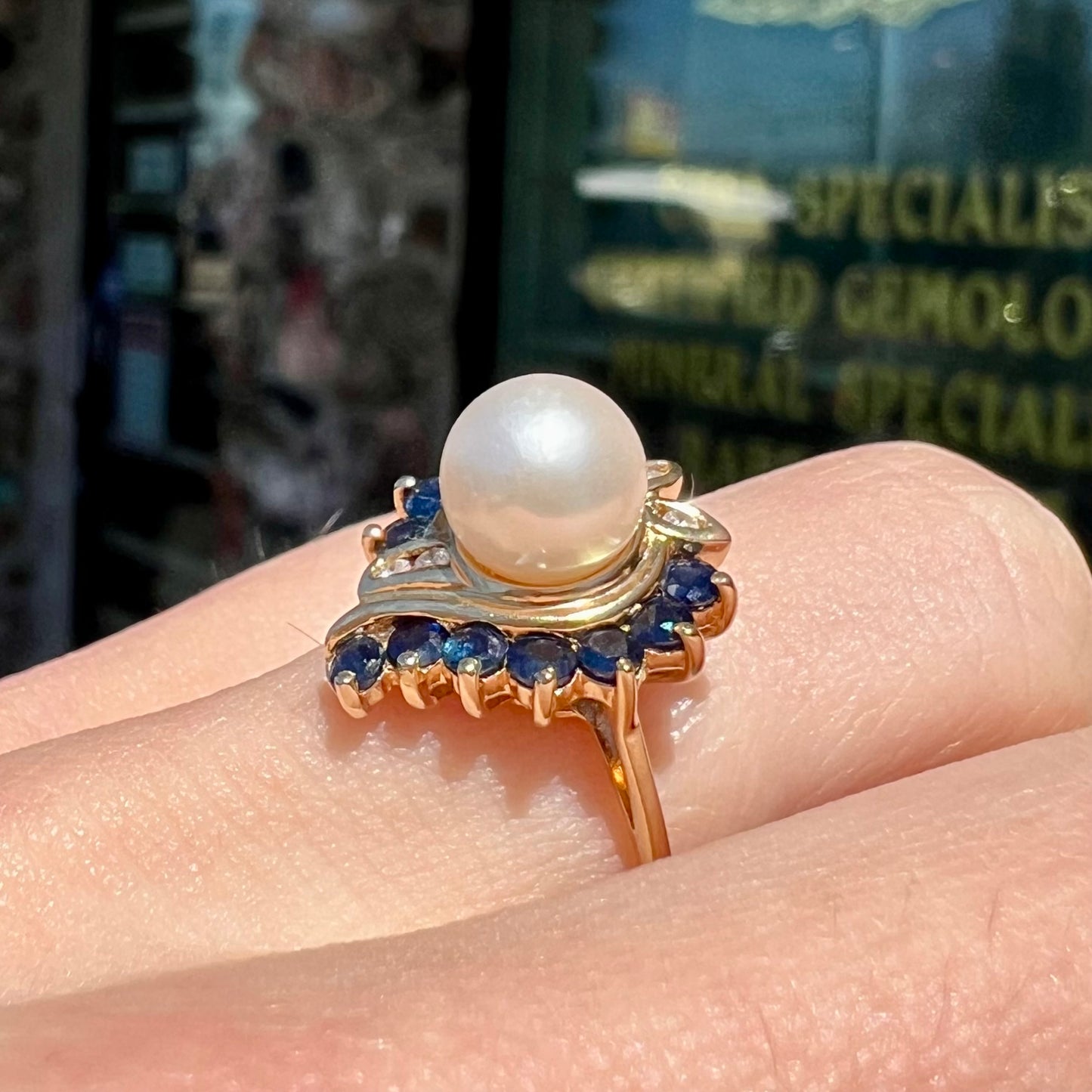 A yellow gold, heart shaped ring set with a freshwater pearl surrounded by blue sapphire and diamond accent stones.