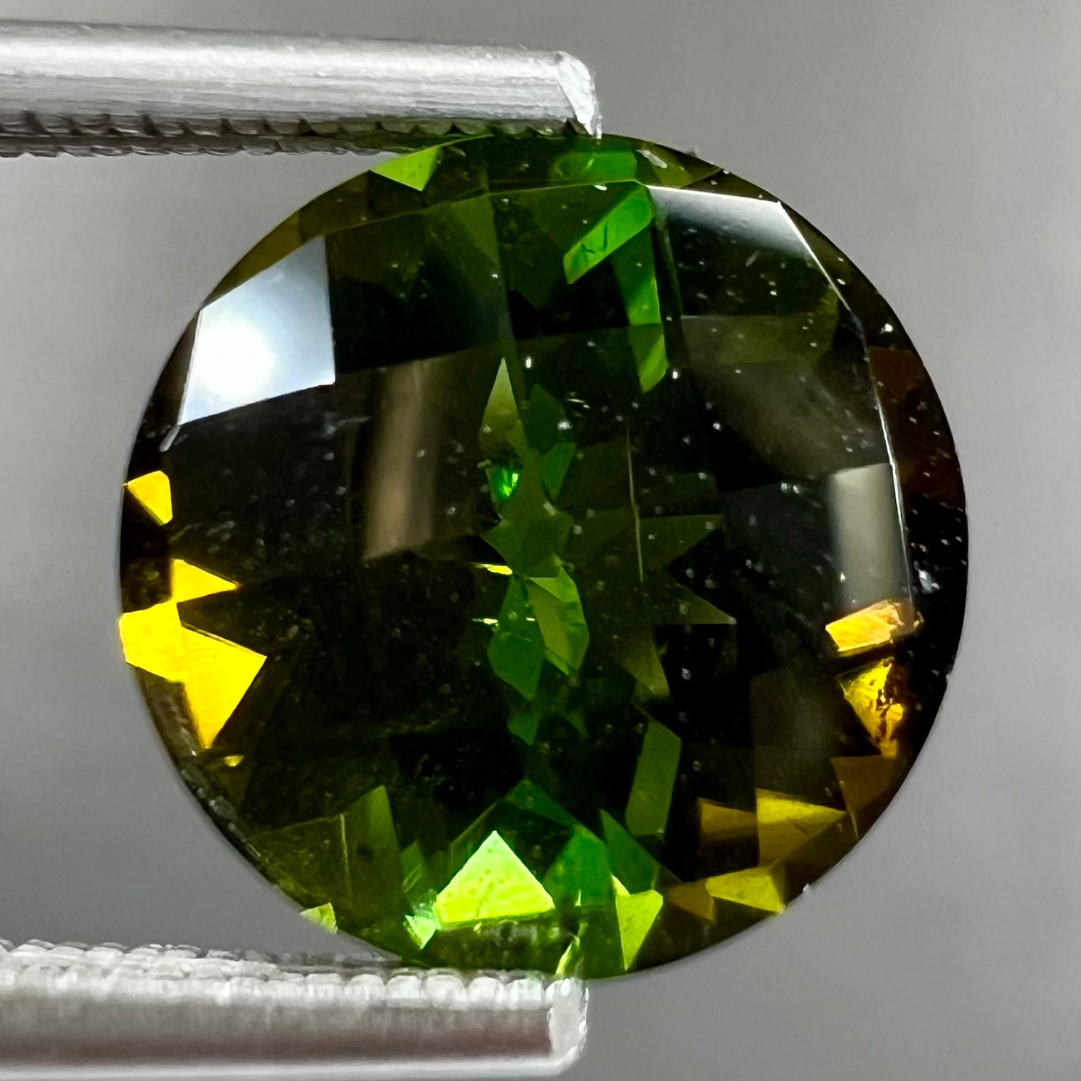 A loose, round checkerboard cut green tourmaline stone with flashes of yellow.