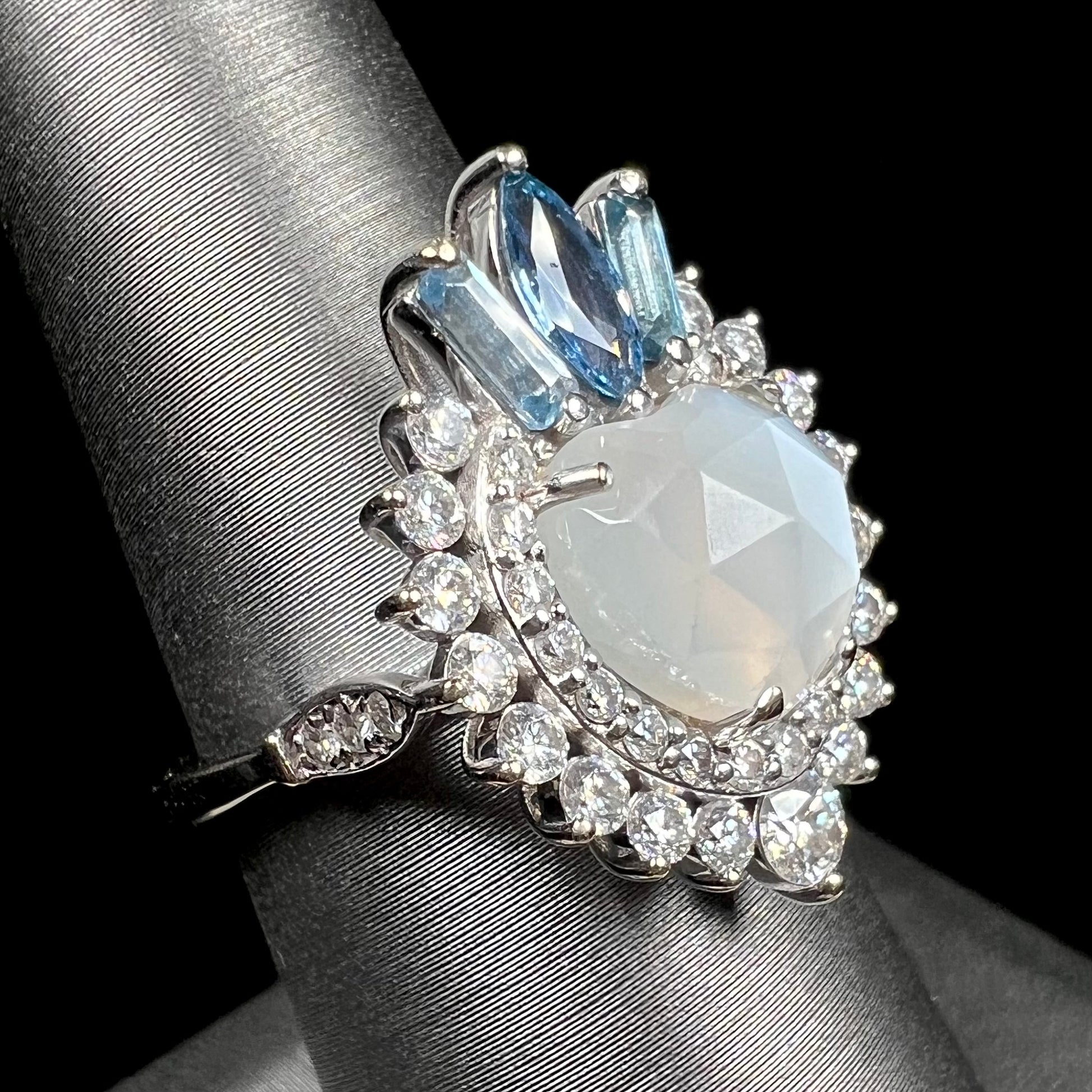 A ladies' white gold ring set with a faceted, heart shaped white moonstone, round moissanites, two aquamarine baguettes, and a marquise cut blue sapphire.