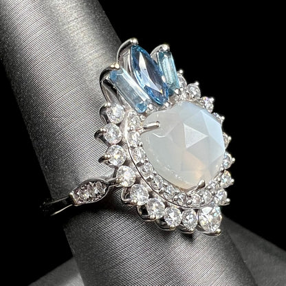 A ladies' white gold ring set with a faceted, heart shaped white moonstone, round moissanites, two aquamarine baguettes, and a marquise cut blue sapphire.