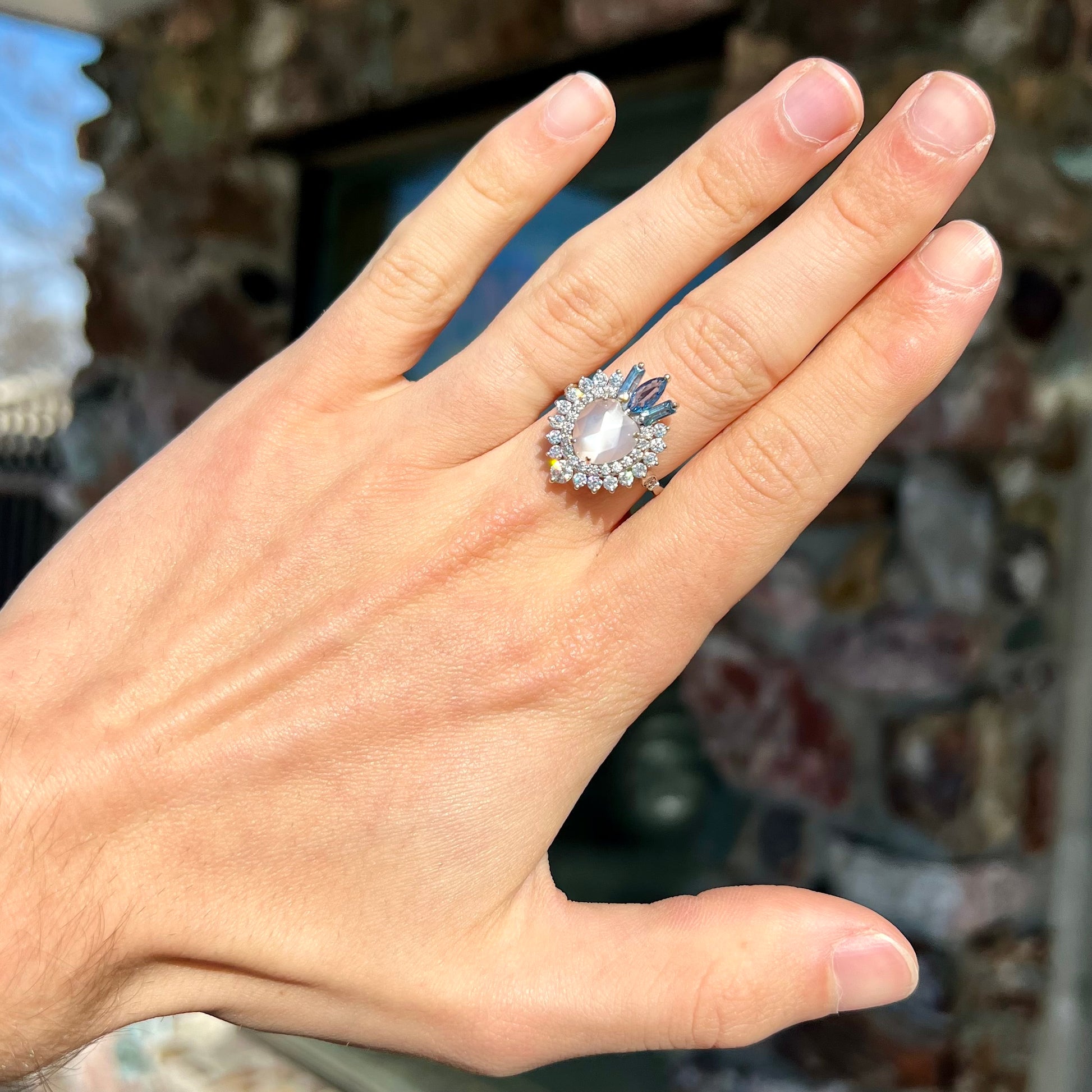 A ladies' white gold ring set with a faceted, heart shaped white moonstone, round moissanites, two aquamarine baguettes, and a marquise cut blue sapphire.