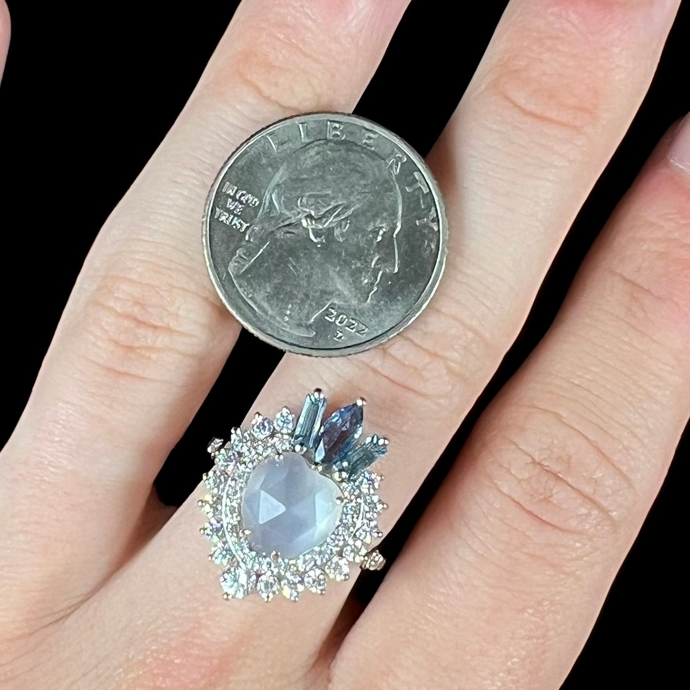 A ladies' white gold ring set with a faceted, heart shaped white moonstone, round moissanites, two aquamarine baguettes, and a marquise cut blue sapphire.