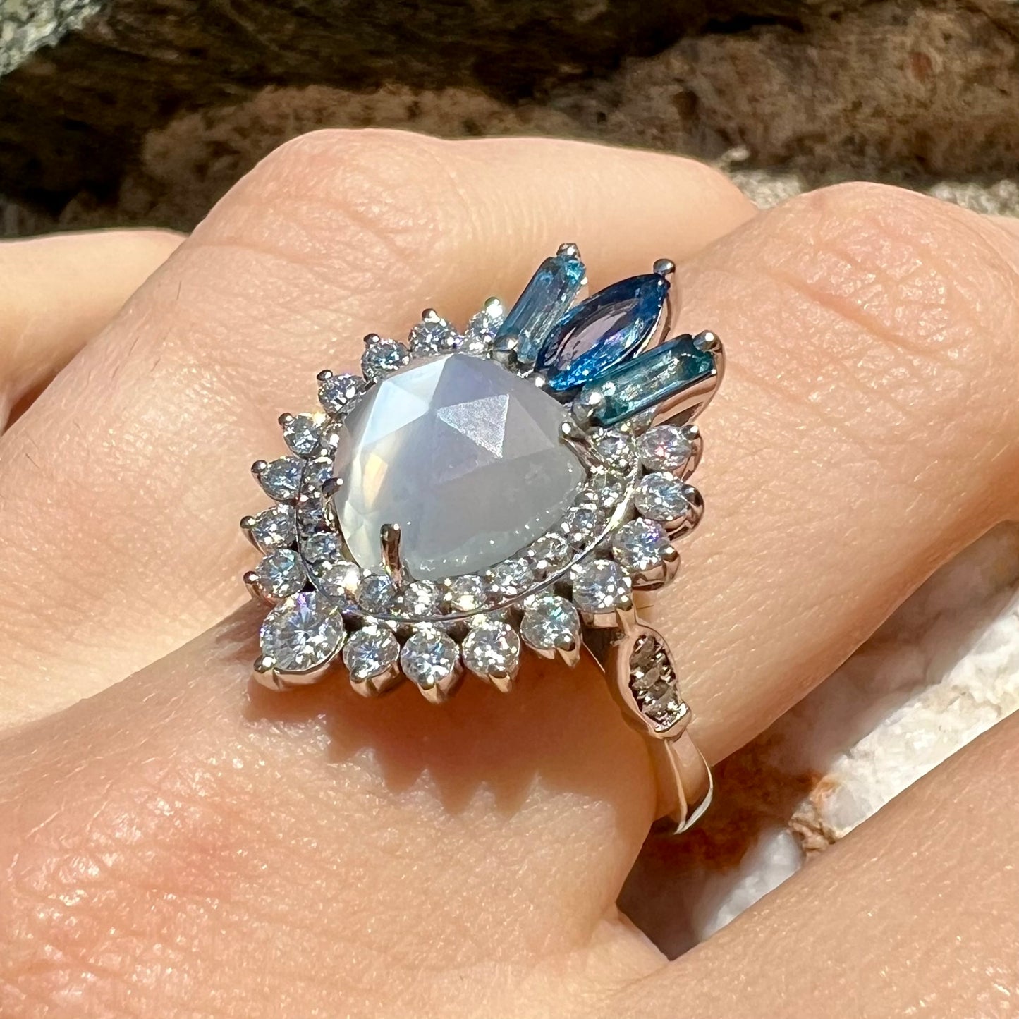 A ladies' white gold ring set with a faceted, heart shaped white moonstone, round moissanites, two aquamarine baguettes, and a marquise cut blue sapphire.