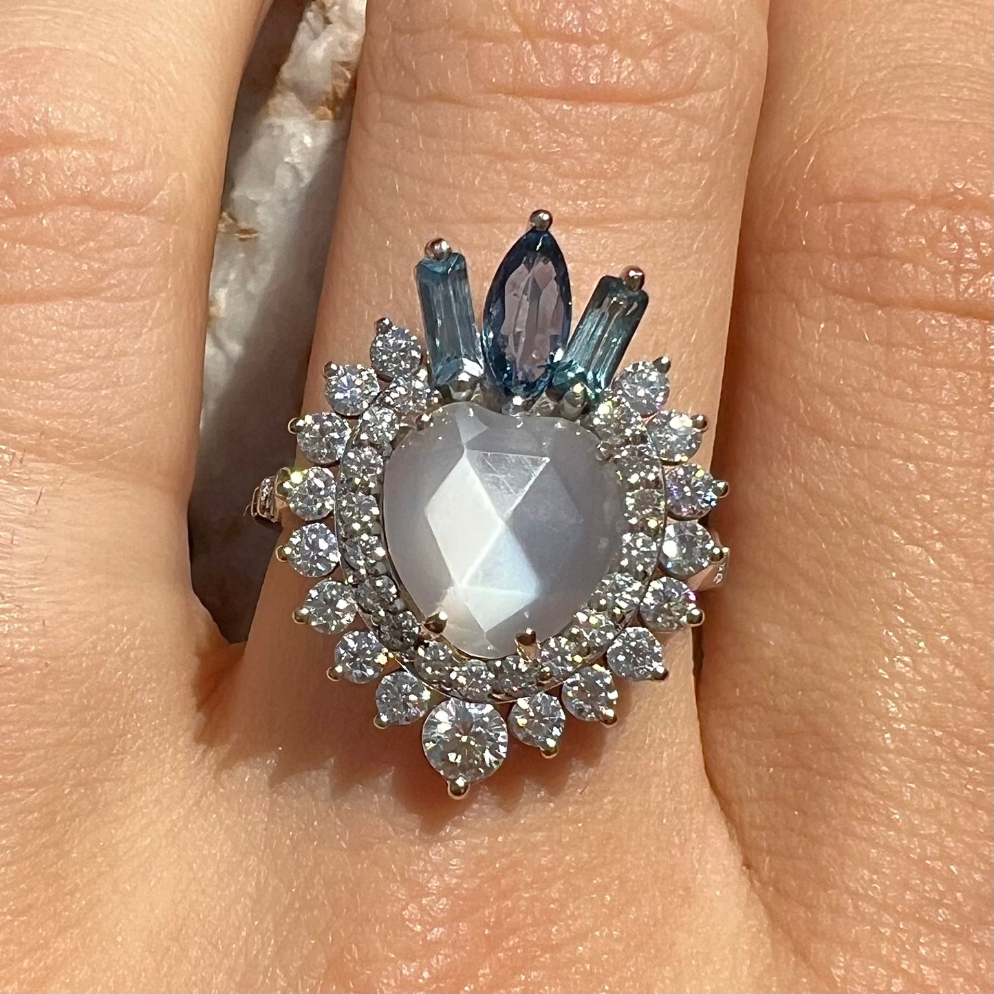 A ladies' white gold ring set with a faceted, heart shaped white moonstone, round moissanites, two aquamarine baguettes, and a marquise cut blue sapphire.