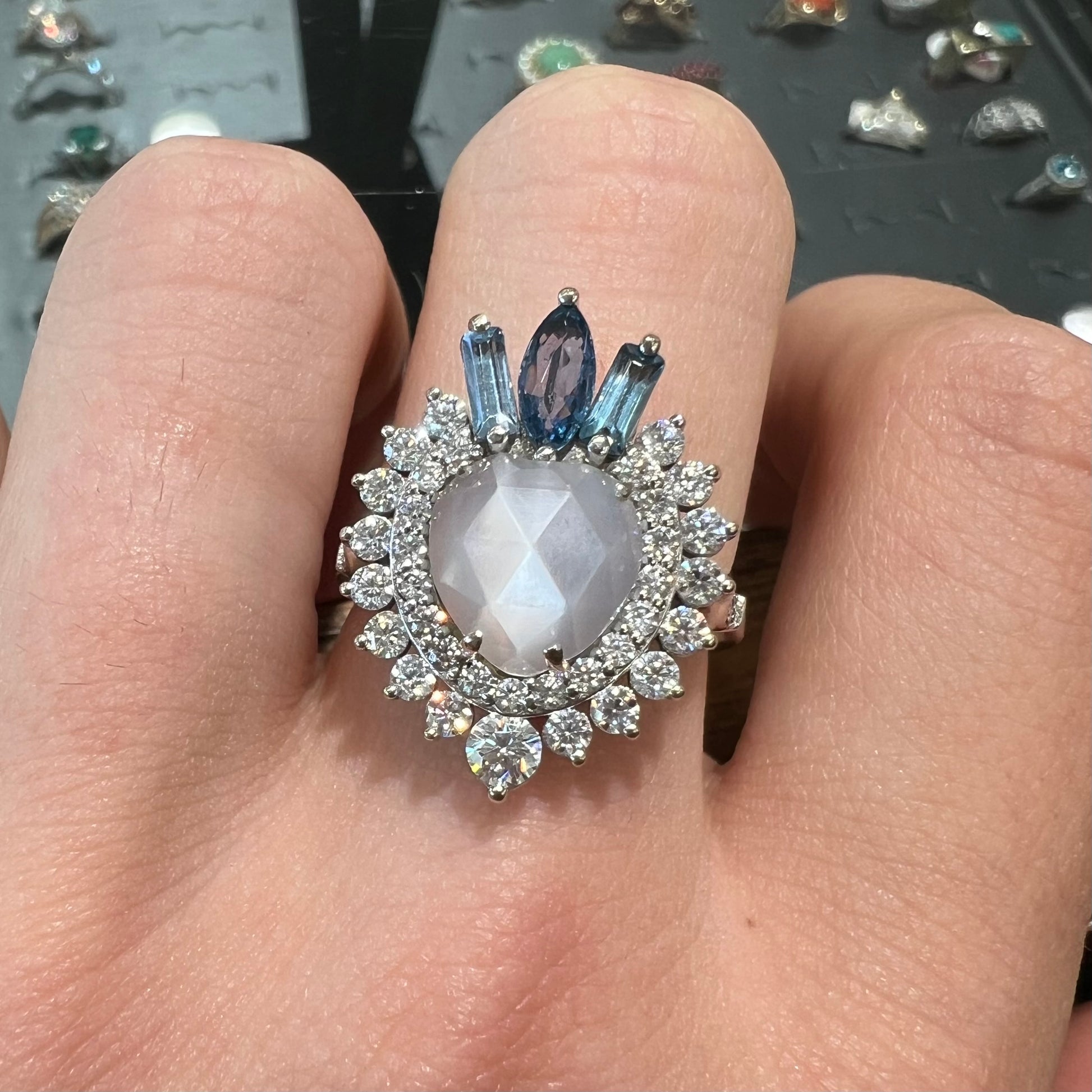 A ladies' white gold ring set with a faceted, heart shaped white moonstone, round moissanites, two aquamarine baguettes, and a marquise cut blue sapphire.