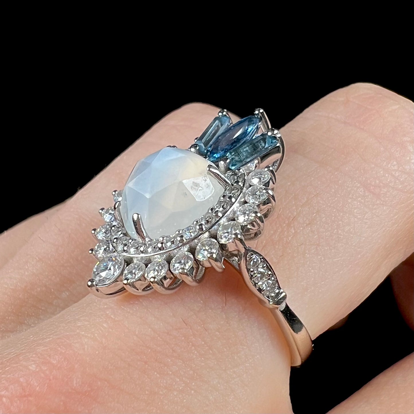 A ladies' white gold ring set with a faceted, heart shaped white moonstone, round moissanites, two aquamarine baguettes, and a marquise cut blue sapphire.