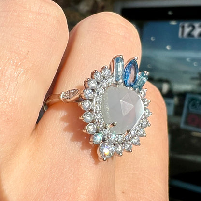 A ladies' white gold ring set with a faceted, heart shaped white moonstone, round moissanites, two aquamarine baguettes, and a marquise cut blue sapphire.