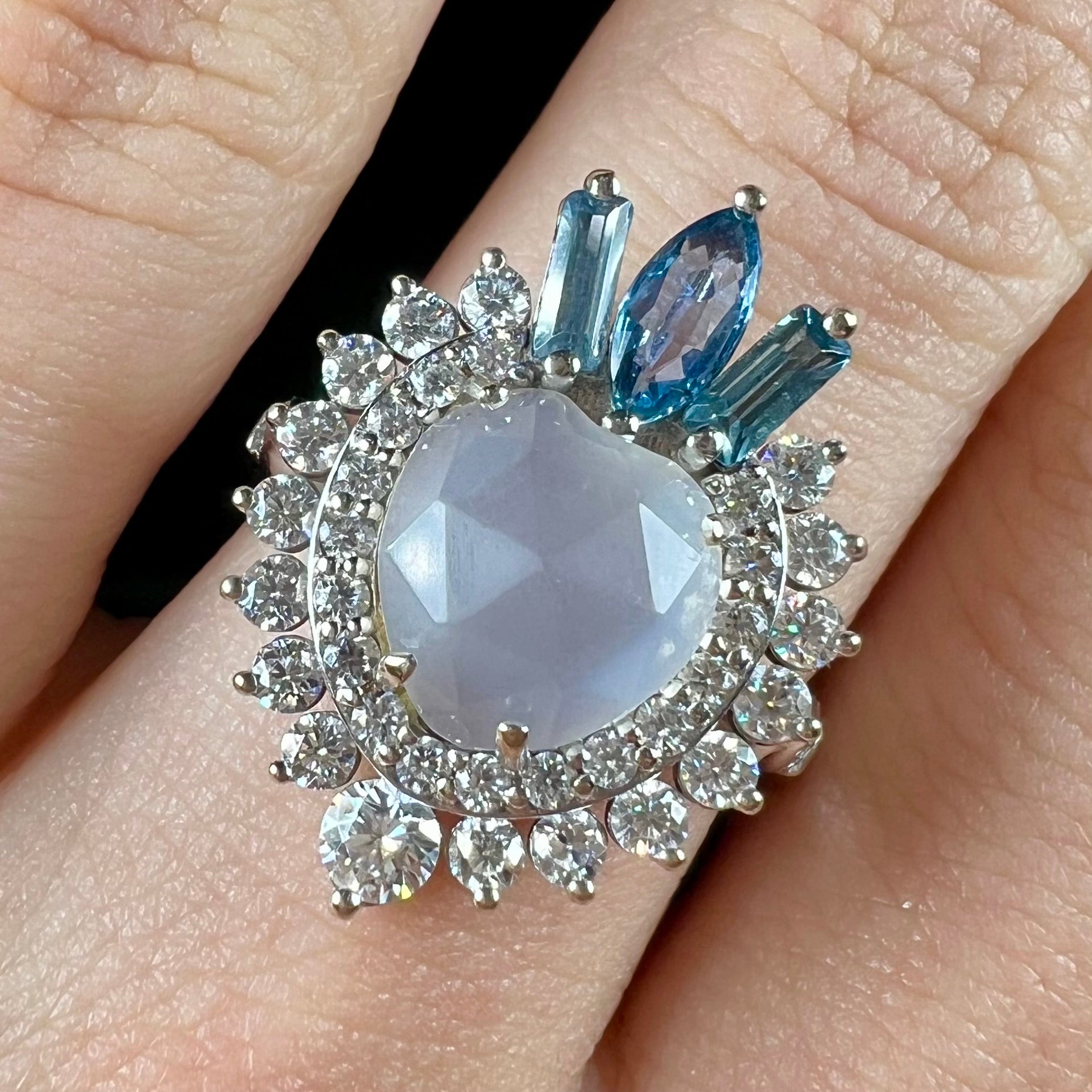 A ladies' white gold ring set with a faceted, heart shaped white moonstone, round moissanites, two aquamarine baguettes, and a marquise cut blue sapphire.