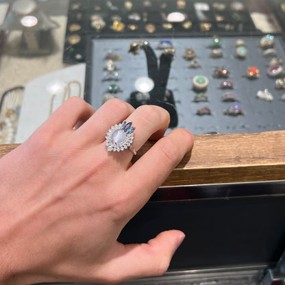 A ladies' white gold ring set with a faceted, heart shaped white moonstone, round moissanites, two aquamarine baguettes, and a marquise cut blue sapphire.