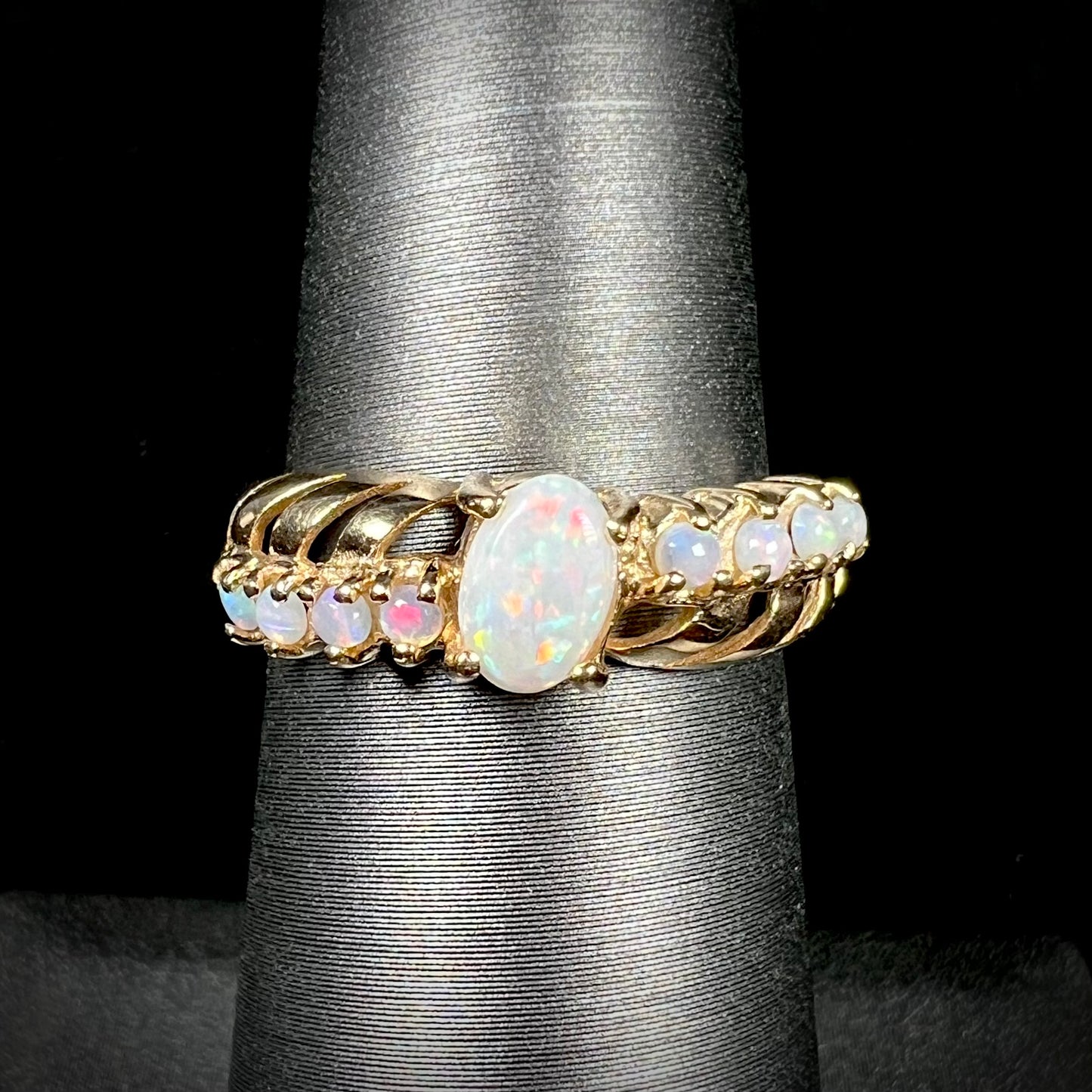 A yellow gold ring featuring two crossing, alternating lines of round cut synthetic opals.  In the middle is a larger oval cut synthetic opal.