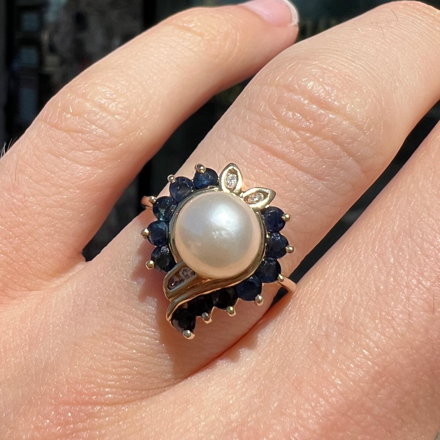 A yellow gold, heart shaped ring set with a freshwater pearl surrounded by blue sapphire and diamond accent stones.