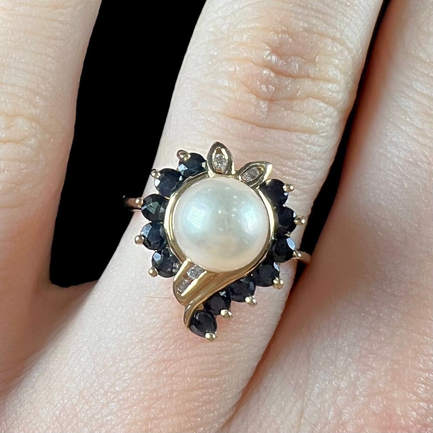 A yellow gold, heart shaped ring set with a freshwater pearl surrounded by blue sapphire and diamond accent stones.