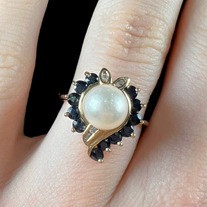 A yellow gold, heart shaped ring set with a freshwater pearl surrounded by blue sapphire and diamond accent stones.