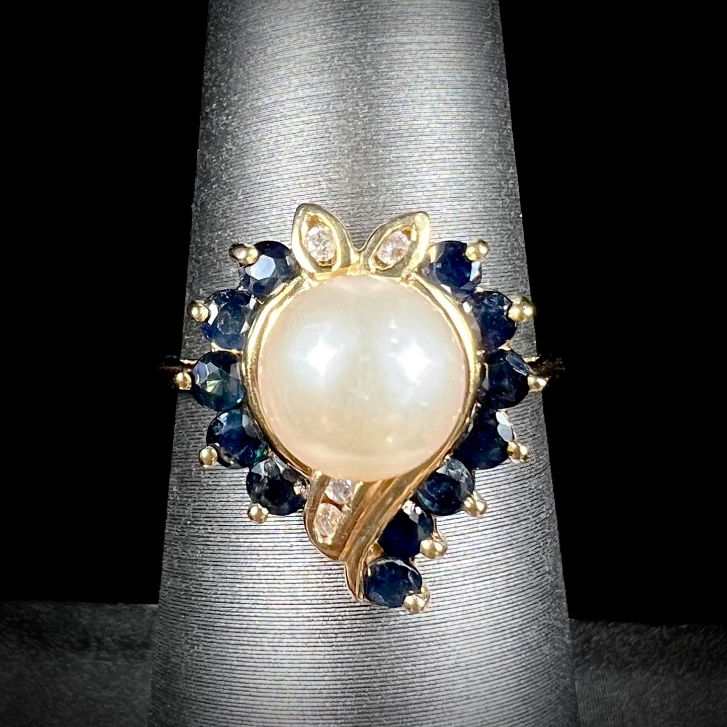 A yellow gold, heart shaped ring set with a freshwater pearl surrounded by blue sapphire and diamond accent stones.