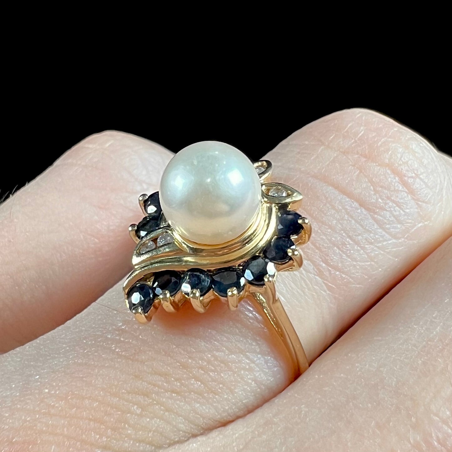 A yellow gold, heart shaped ring set with a freshwater pearl surrounded by blue sapphire and diamond accent stones.