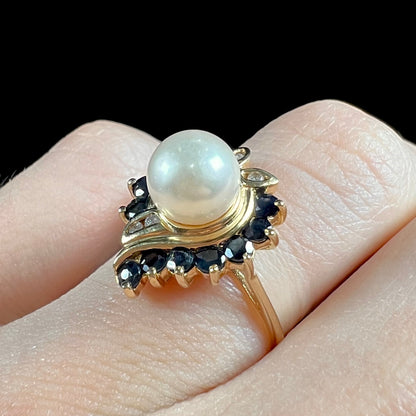 A yellow gold, heart shaped ring set with a freshwater pearl surrounded by blue sapphire and diamond accent stones.