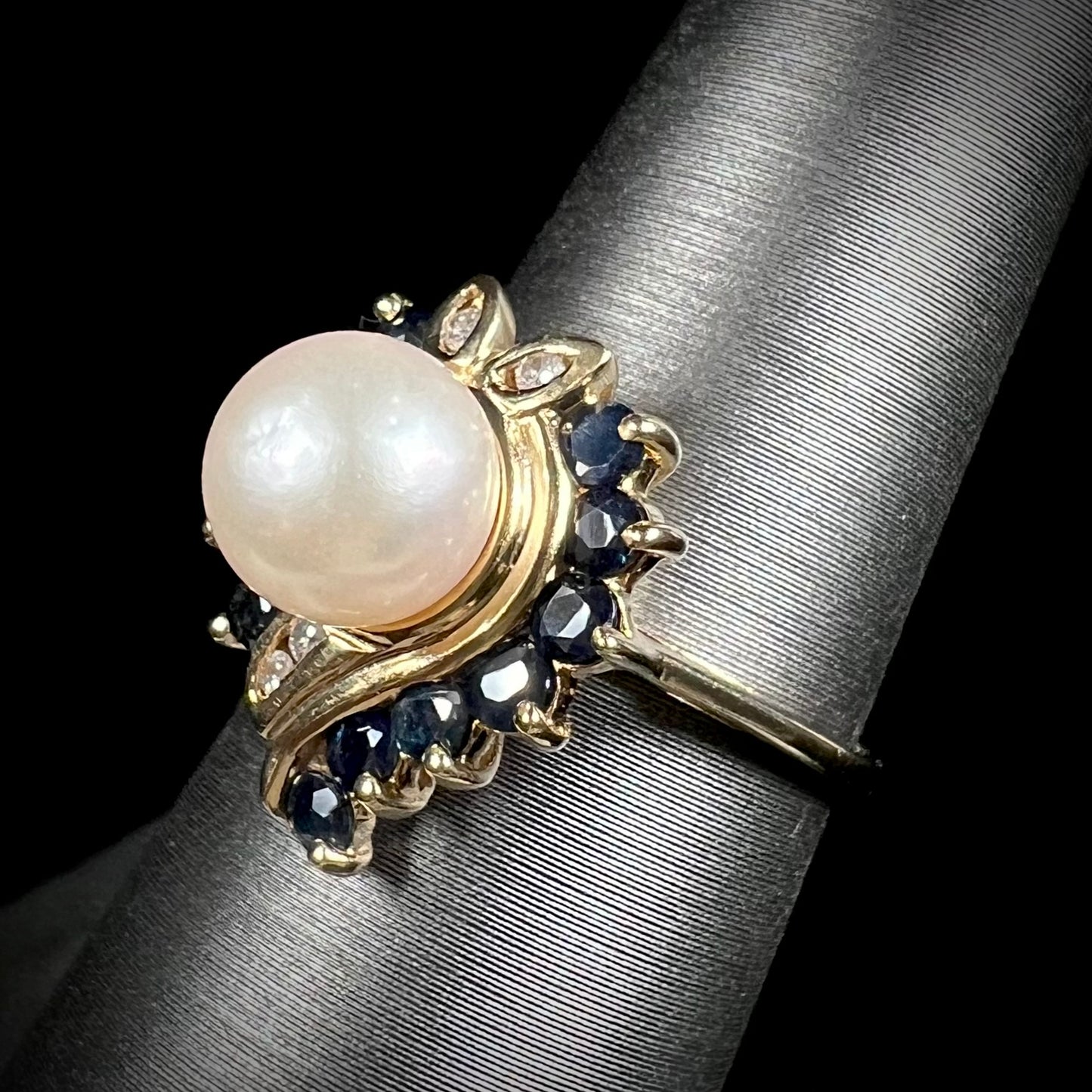 A yellow gold, heart shaped ring set with a freshwater pearl surrounded by blue sapphire and diamond accent stones.