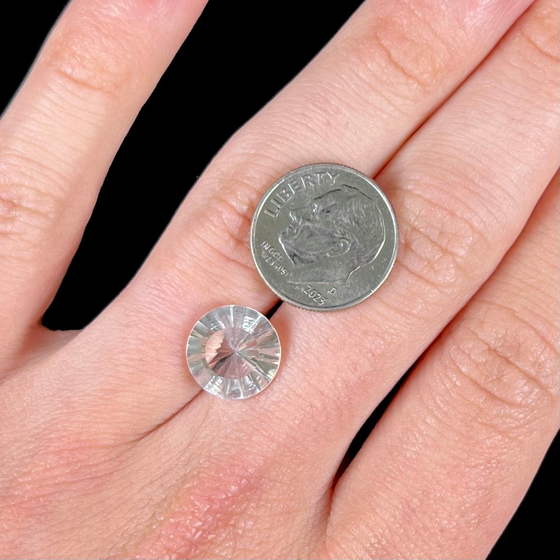 A loose, round cut petalite gemstone.  The faceted stone is colorless with a black surface inclusion.