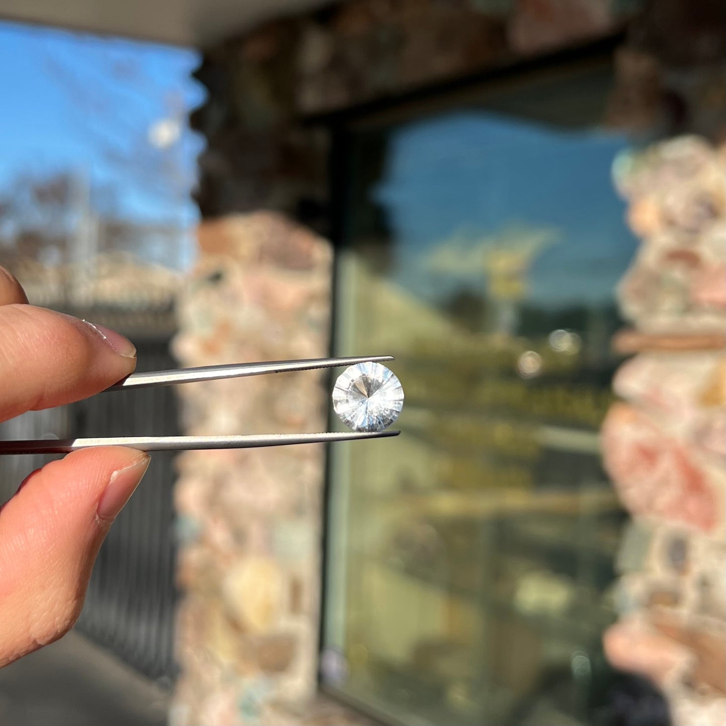 A loose, round cut petalite gemstone.  The faceted stone is colorless with a black surface inclusion.