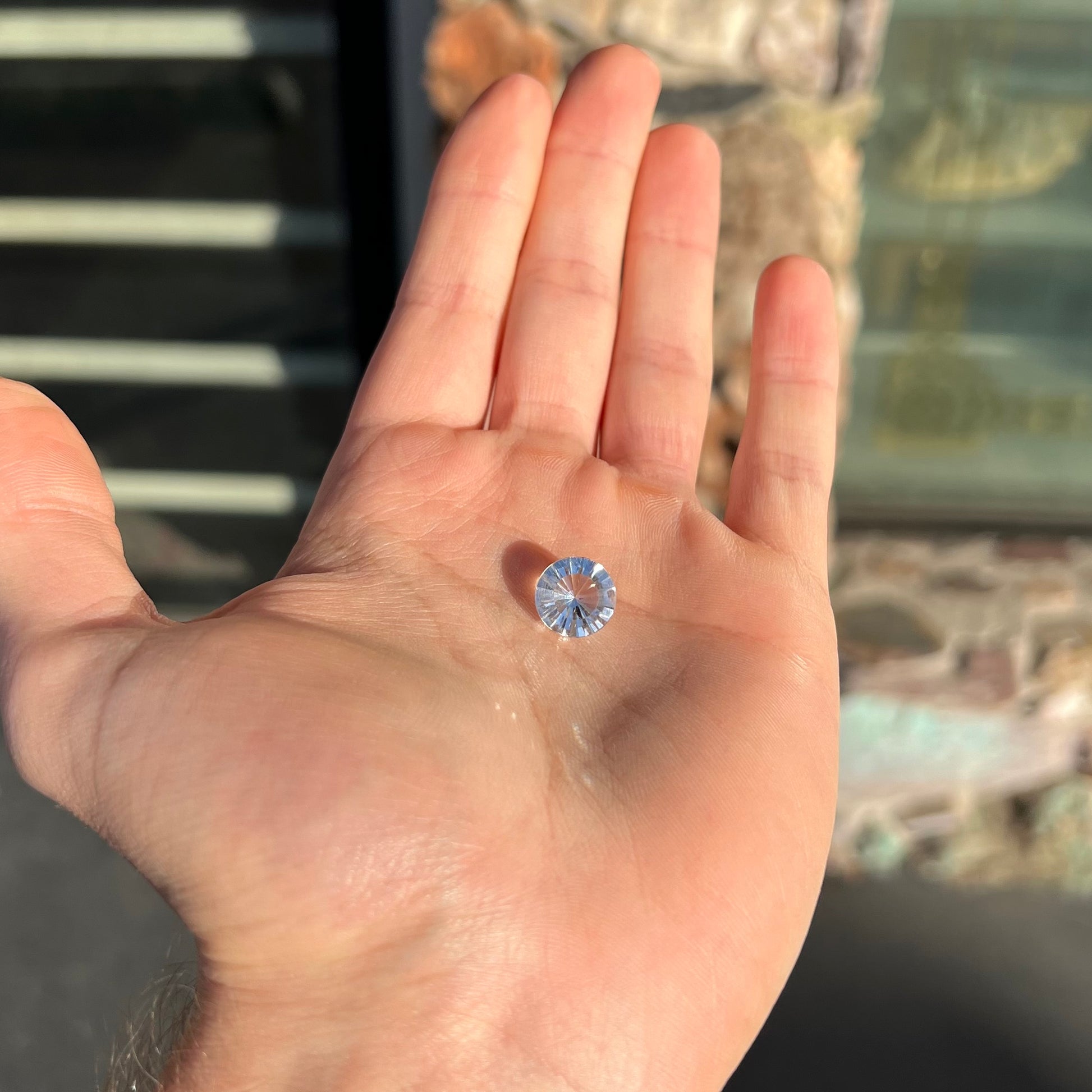 A loose, round cut petalite gemstone.  The faceted stone is colorless with a black surface inclusion.