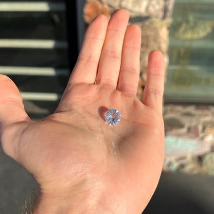 A loose, round cut petalite gemstone.  The faceted stone is colorless with a black surface inclusion.