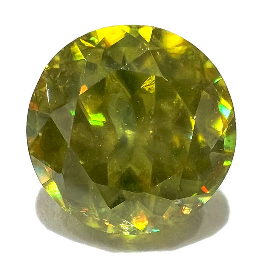 A loose, round brilliant cut sphene gemstone.  The stone is green with orange, green, blue, and yellow flashes.