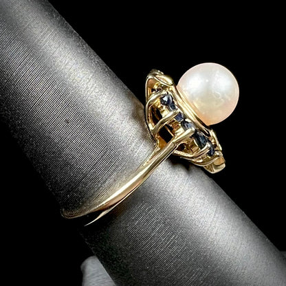 A yellow gold, heart shaped ring set with a freshwater pearl surrounded by blue sapphire and diamond accent stones.