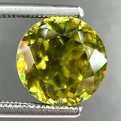 A loose, round brilliant cut sphene gemstone.  The stone is green with orange, green, blue, and yellow flashes.