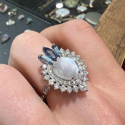 A ladies' white gold ring set with a faceted, heart shaped white moonstone, round moissanites, two aquamarine baguettes, and a marquise cut blue sapphire.