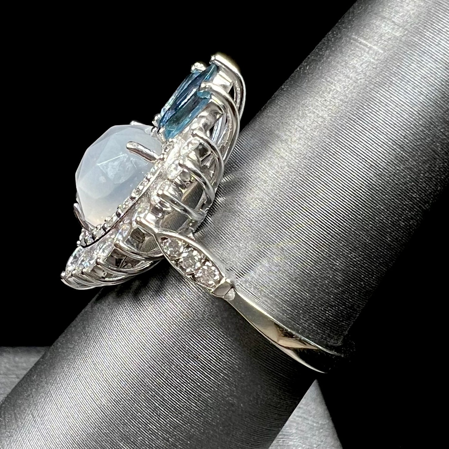 A ladies' white gold ring set with a faceted, heart shaped white moonstone, round moissanites, two aquamarine baguettes, and a marquise cut blue sapphire.