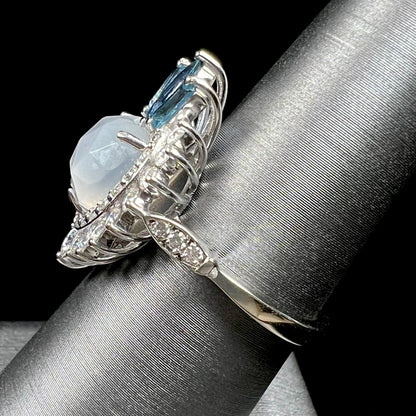 A ladies' white gold ring set with a faceted, heart shaped white moonstone, round moissanites, two aquamarine baguettes, and a marquise cut blue sapphire.