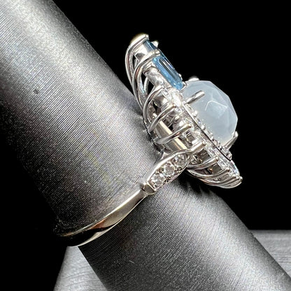 A ladies' white gold ring set with a faceted, heart shaped white moonstone, round moissanites, two aquamarine baguettes, and a marquise cut blue sapphire.