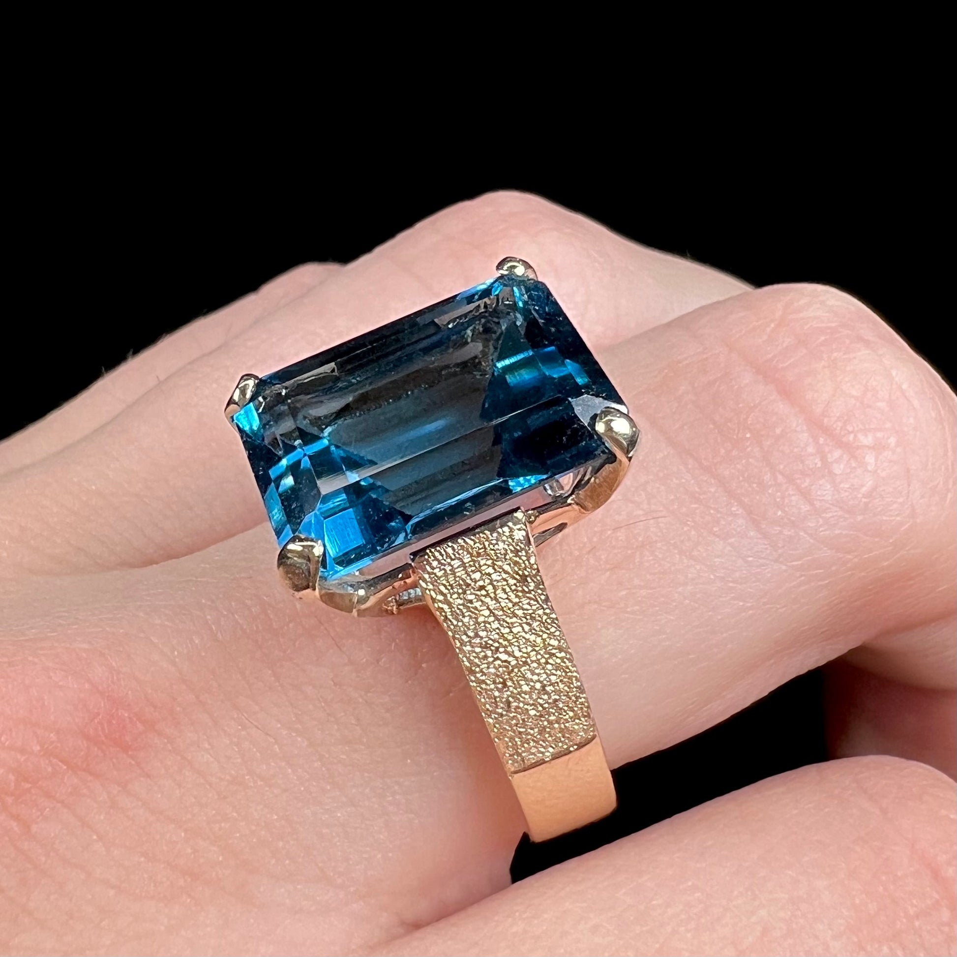 A ladies' yellow gold London blue topaz solitaire ring.  The ring is textured, and the topaz is emerald cut.