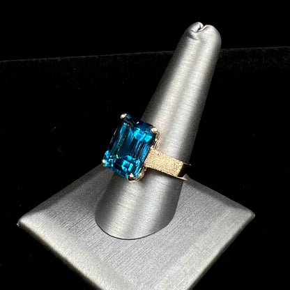 A ladies' yellow gold London blue topaz solitaire ring.  The ring is textured, and the topaz is emerald cut.