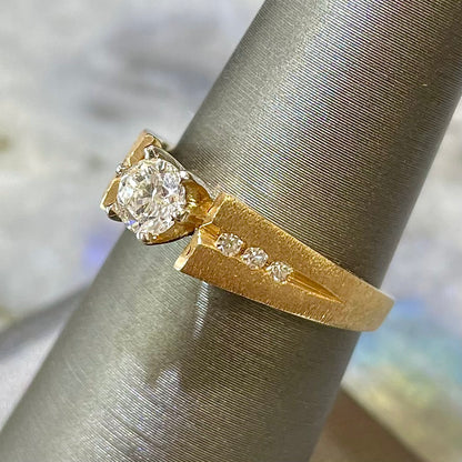 A diamond ring that features one Standard Round Brilliant Cut diamond set in a six prong head with three side diamonds channel set along side it in a 14kt yellow gold satin/matte finish mounting.  The piece is on a ring holder.