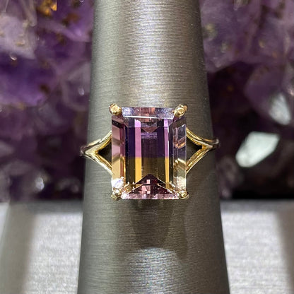 Solitaire 4 prong set ametrine quartz ring in 14kt yellow gold.  Split shank ring.  The ametrine displays a near perfect color separation with purple on the top and golden yellow on the bottom.  The ring is on a stand against a purple amethyst crystal background.